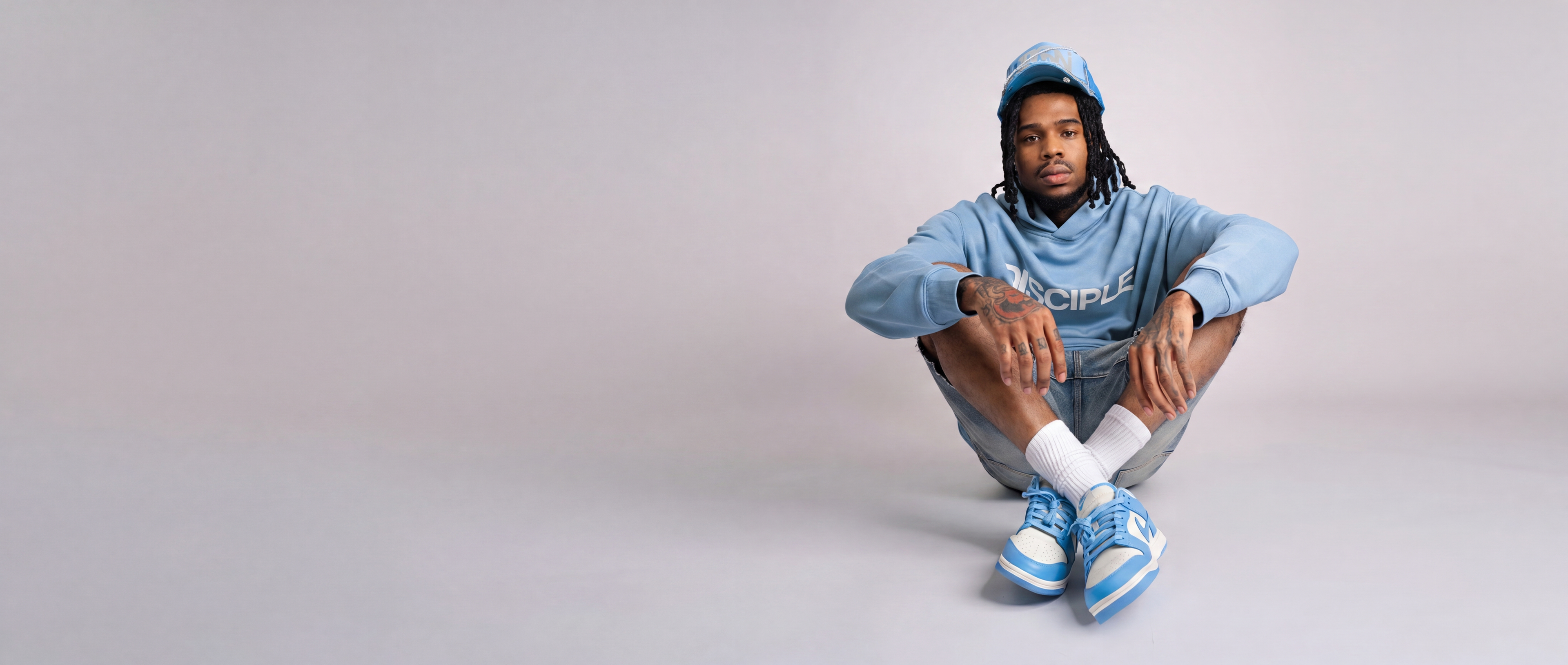 Man seated in light-blue DISCIPLE hoodie, cap, shorts and sneakers, wide studio banner